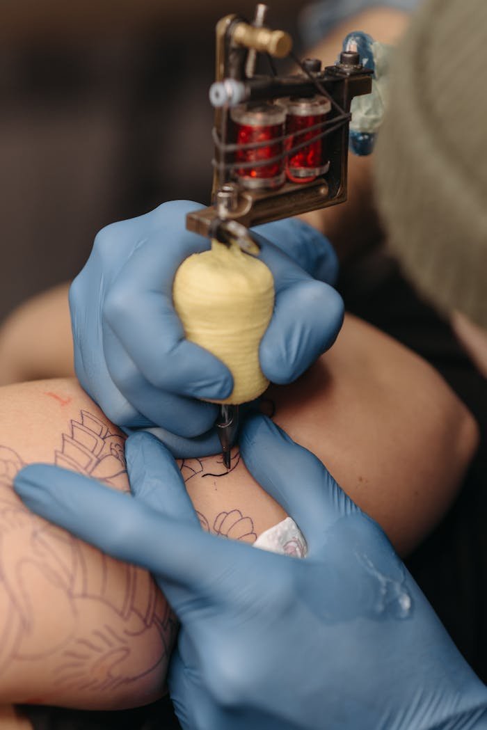 about-me-02 Close-up of a tattoo artist using a tattoo machine on an arm, wearing blue latex gloves for precision.