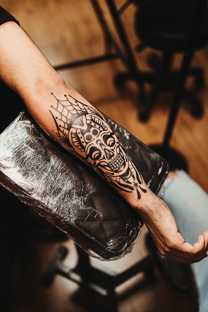 social-feed-img-01 Close-up of a detailed skull tattoo on a mans forearm in a tattoo studio setting.